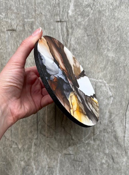 A round, earthy coloured abstract art piece, done in a marbled stone effect, held in hand against a grey stone wall.