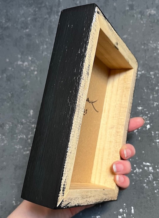 Hand holding a cradled panel with a black exterior and natural wood interior in a grey interior room.