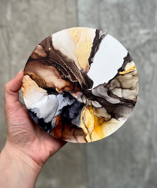 A round, marble effect abstract wall art painting featuring earthy colours, held in a hand above a neutral stone background.