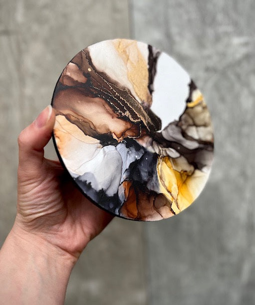 Round marbleised abstract wall art painting in earthy colours, held in a hand above a grey stone background.