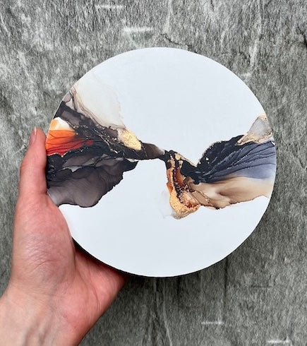 Gothic home decor of abstract autumn wall art featuring black, orange, brown and gold colours unfurling like wings on a white, circle panel, held before a stone wall.