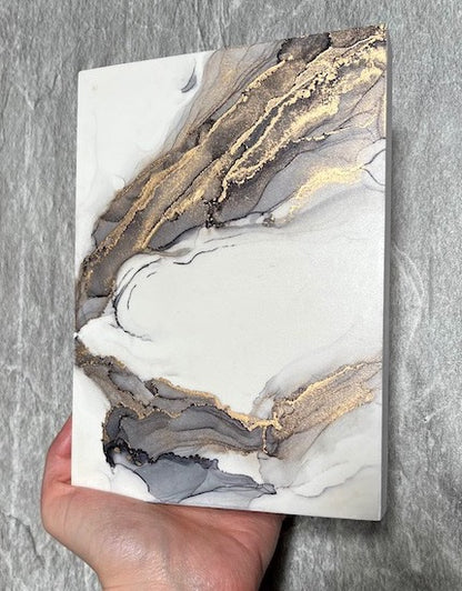 Abstract wall art of fantasy gothic wings, hand painted in grey and gold colours with a marble stone technique, held in a hand above a stone backdrop.