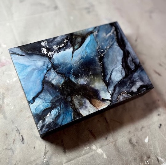 Abstract art conveying the feel of an ocean through a marbled blue and black stone effect. This hand painted blue abstract art rests on a grey background.