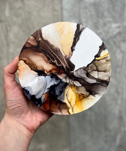 A round, marble effect abstract wall art painting featuring earthy colours, held in a hand above a neutral stone background.