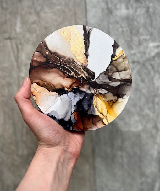 Round, abstract wall art akin to marble stone featuring earthy colours, held in a hand above a grey background.