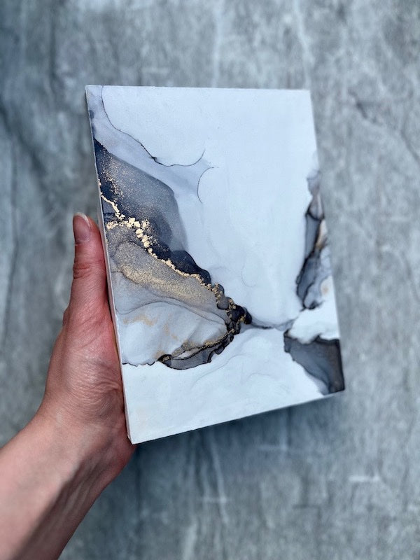 An original painting for sale featuring ethereal gothic wings. A 5x7" grey and gold wall art held in a hand above a stone wall.