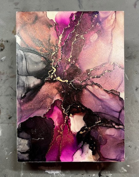 Abstract space art where shades of purple, pink, black, copper and gold collide in a moody expression of starlight and lightning. This abstract art piece rests on a grey surface.